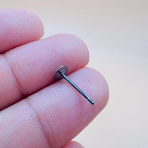 Pu&ograve; includere: Un singolo perno per orecchino color argento con un disco piatto e rotondo e un perno sottile e dritto. Il perno dell'orecchino &egrave; tenuto tra le dita su uno sfondo beige chiaro. &Egrave; un componente di gioielleria.