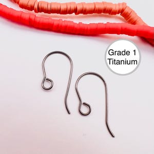 Può includere: Due ganci per orecchini in titanio di grado 1 color argento. I ganci hanno un design curvo con un piccolo anello nella parte superiore e un fondo più lungo e curvo. Lo sfondo è bianco con perline rosse e arancioni.