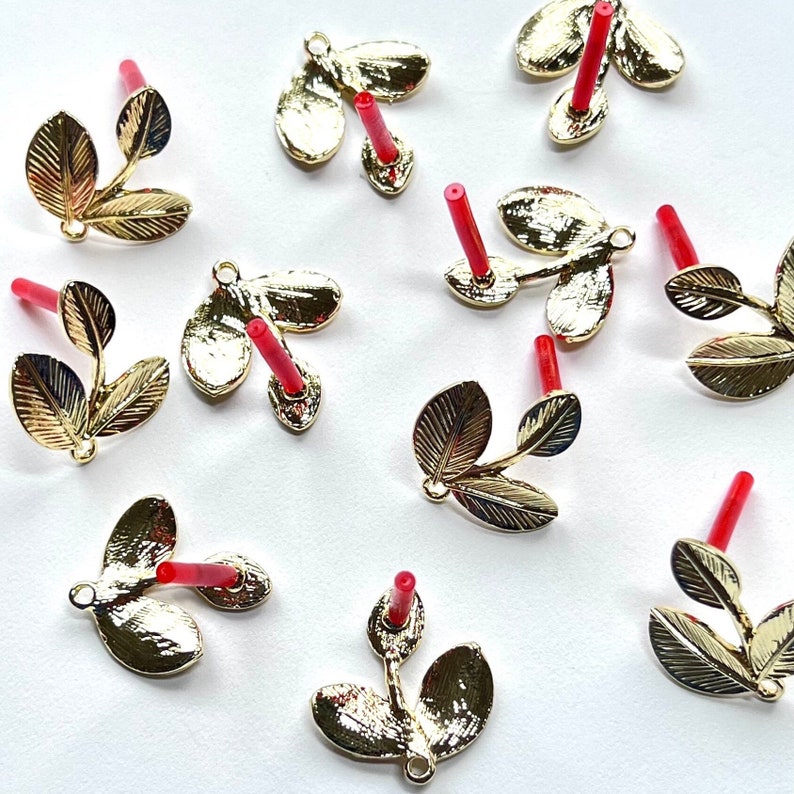 May include: Ten gold leaf charms with red stems. Each charm features three leaves and a loop for attaching to a jewelry piece.