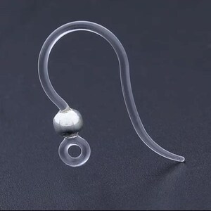 May include: Clear, flexible earring hook with a silver-colored bead and a small loop at the bottom. The hook curves upward and then back down, designed to fit through a pierced ear. The earring hook is made of a transparent material.