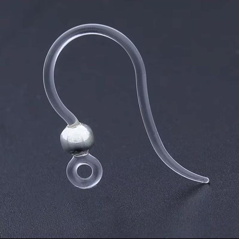 May include: Clear, flexible earring hook with a silver-colored bead and a small loop at the bottom. The hook curves upward and then back down, designed to fit through a pierced ear. The earring hook is made of a transparent material.