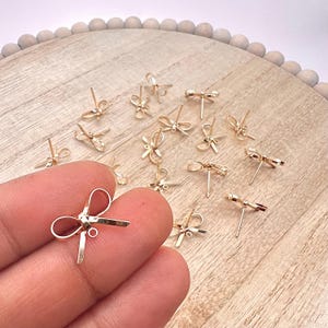May include: A group of gold-toned bow-shaped earring findings on a wooden surface. The findings are small and delicate, with a loop at the top for attaching a earring.