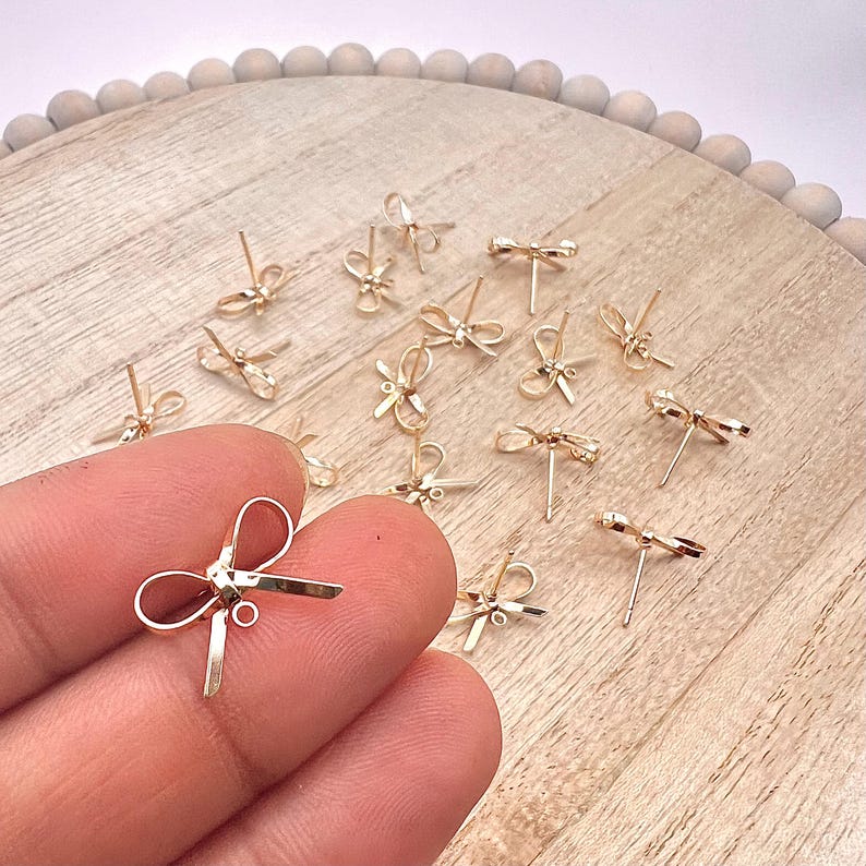 May include: A group of gold-toned bow-shaped earring findings on a wooden surface. The findings are small and delicate, with a loop at the top for attaching a earring.