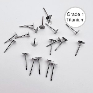 May include: A collection of Grade 1 Titanium earring posts scattered on a white surface. The posts have a silver color with a flat, round disc at the top and a straight post extending downwards. The text "Grade 1 Titanium" is in a white circle.