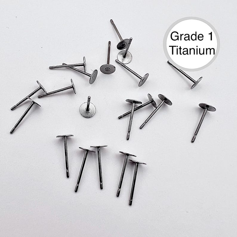 Pu&ograve; includere: Una collezione di perni per orecchini in titanio di grado 1 sparsi su una superficie bianca. I perni sono di colore argento con un disco piatto e rotondo nella parte superiore e un perno dritto che si estende verso il basso. Il testo "Grade 1 Titanium" &egrave; in un cerchio bianco.