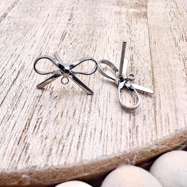 May include: A pair of silver bow-shaped earrings with a post and a loop for attaching charms. The earrings are on a wooden surface.