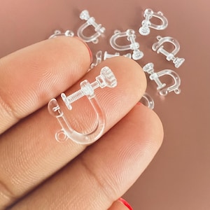 May include: Clear plastic screw-on earring findings. The findings have a U-shaped design with a screw mechanism for secure attachment. Several pieces are scattered, with one in focus, showcasing the details of the design. The findings are small and lightweight.