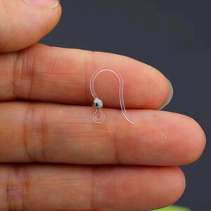 May include: A clear, plastic earring hook with a small, silver-colored bead. The hook is held between fingers, showcasing its delicate design and simple construction. The earring hook is designed for pierced ears.