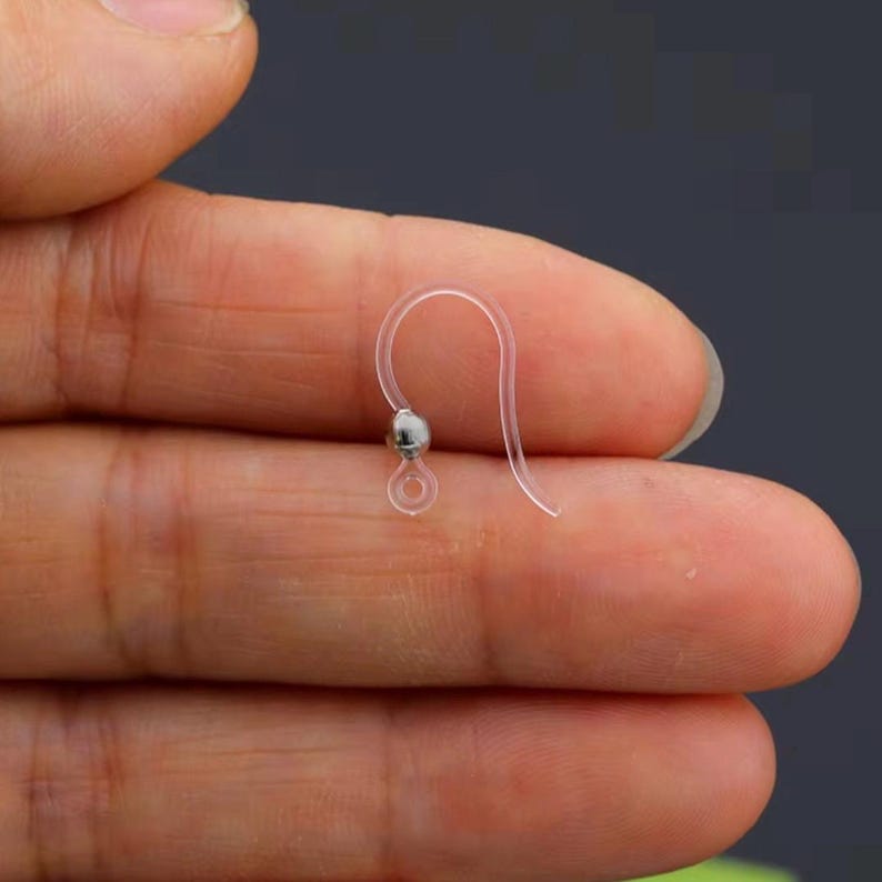May include: A clear, plastic earring hook with a small, silver-colored bead. The hook is held between fingers, showcasing its delicate design and simple construction. The earring hook is designed for pierced ears.