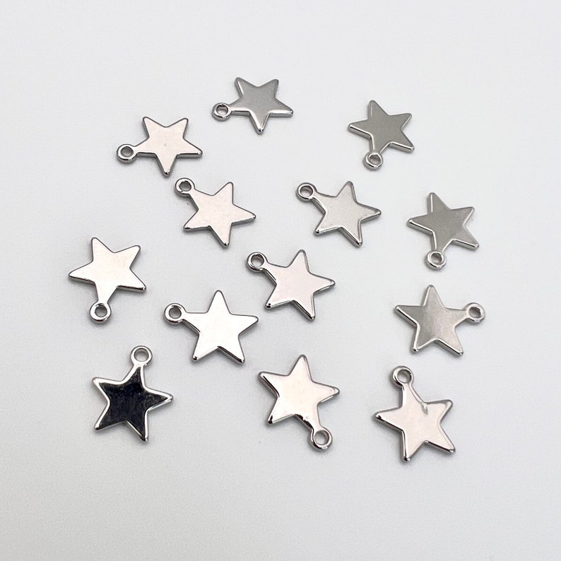 K&ouml;nnte beinhalten: Eine Sammlung von 14 silbernen sternf&ouml;rmigen Charms mit einer Schlaufe an jeder Seite. Die Charms sind auf einer wei&szlig;en Oberfl&auml;che angeordnet.