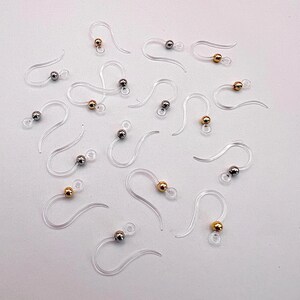 May include: A collection of clear, plastic earring hooks with small, metallic beads. Some beads are gold-colored, and others are silver-colored. The hooks are arranged on a white surface, showcasing their delicate design and functionality for jewelry making.