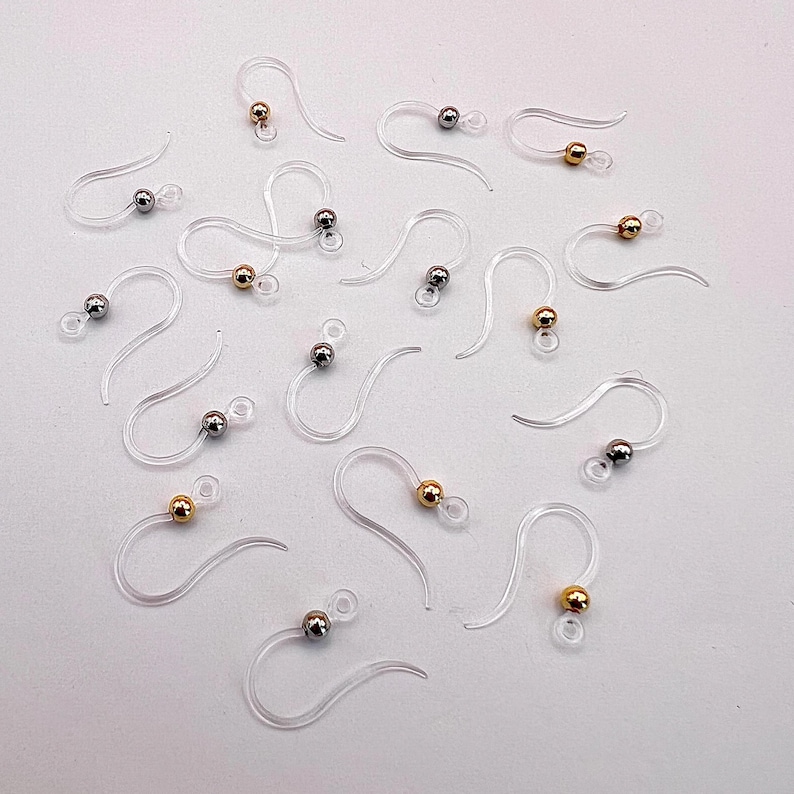 May include: A collection of clear, plastic earring hooks with small, metallic beads. Some beads are gold-colored, and others are silver-colored. The hooks are arranged on a white surface, showcasing their delicate design and functionality for jewelry making.