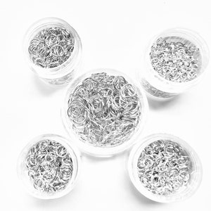 Puede incluir: Cinco frascos de vidrio transparente llenos de anillas de salto plateadas. Los frascos están dispuestos en forma de estrella sobre una superficie blanca.