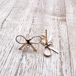 May include: Two gold-toned metal earring findings in the shape of a bow. The findings have a post and a loop for attaching an earring.