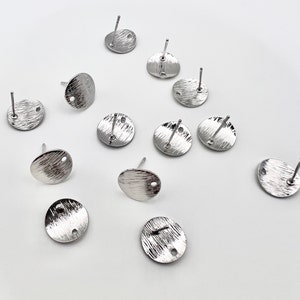 May include: Twelve silver earring blanks with a textured finish and a small hole in the center. The blanks are round and have a post on the back.