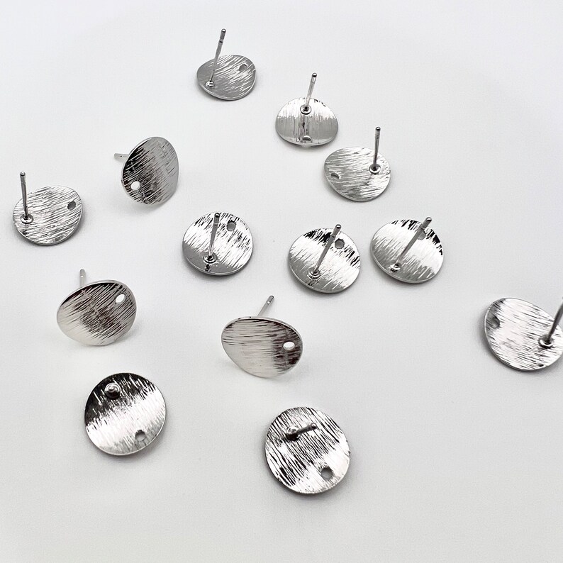 May include: Twelve silver earring blanks with a textured finish and a small hole in the center. The blanks are round and have a post on the back.