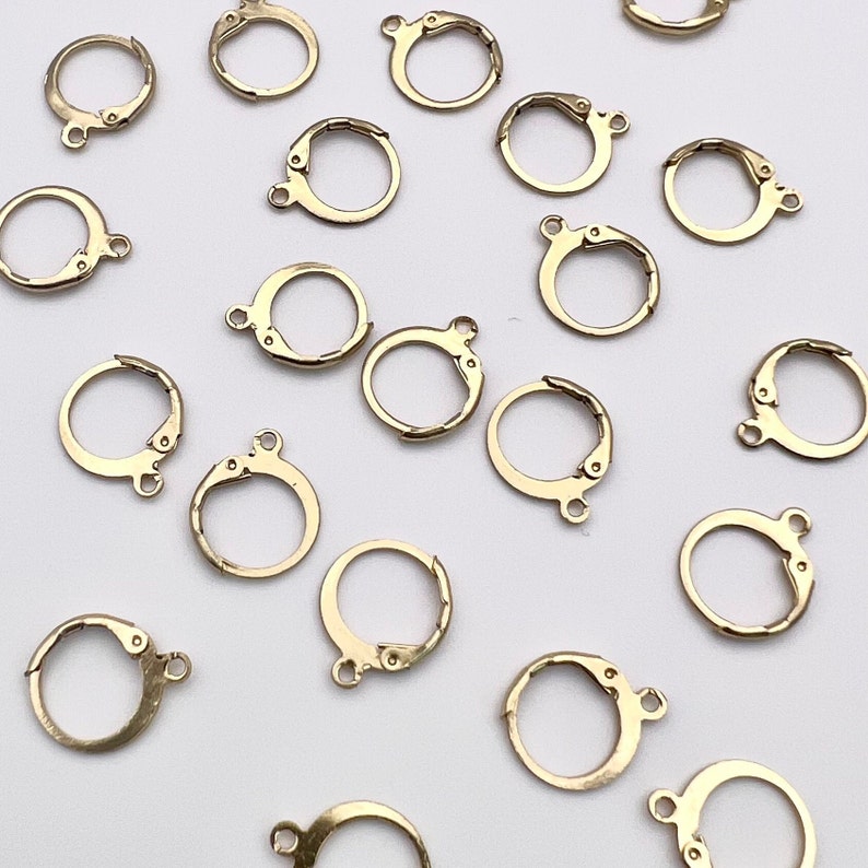 May include: A collection of gold-toned earring clasps, also known as earring findings, are scattered on a white surface. These clasps are used to attach earrings to ear wires or posts.