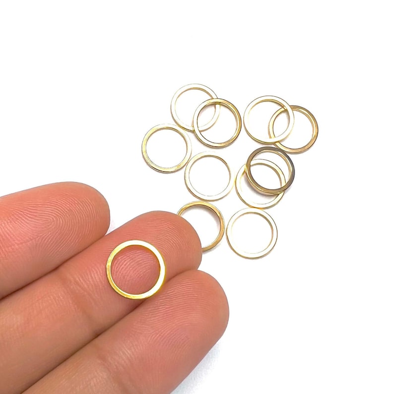 May include: A close-up of a hand holding a gold jump ring. There are several more gold jump rings scattered on a white surface.