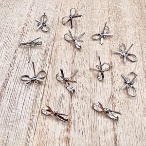May include: A collection of silver-toned bow-shaped earring findings. The findings are arranged on a light wood surface.