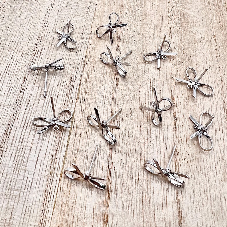 May include: A collection of silver-toned bow-shaped earring findings. The findings are arranged on a light wood surface.