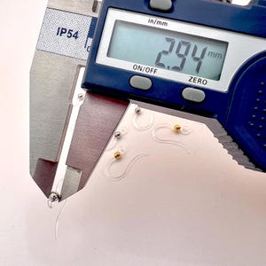 May include: Close-up of a digital caliper measuring a clear plastic earring hook. The digital display reads 2.94 mm. Several clear earring hooks with silver and gold-colored beads are visible.