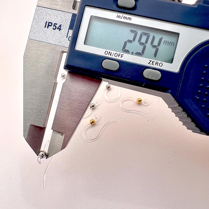 May include: Close-up of a digital caliper measuring a clear plastic earring hook. The digital display reads 2.94 mm. Several clear earring hooks with silver and gold-colored beads are visible.