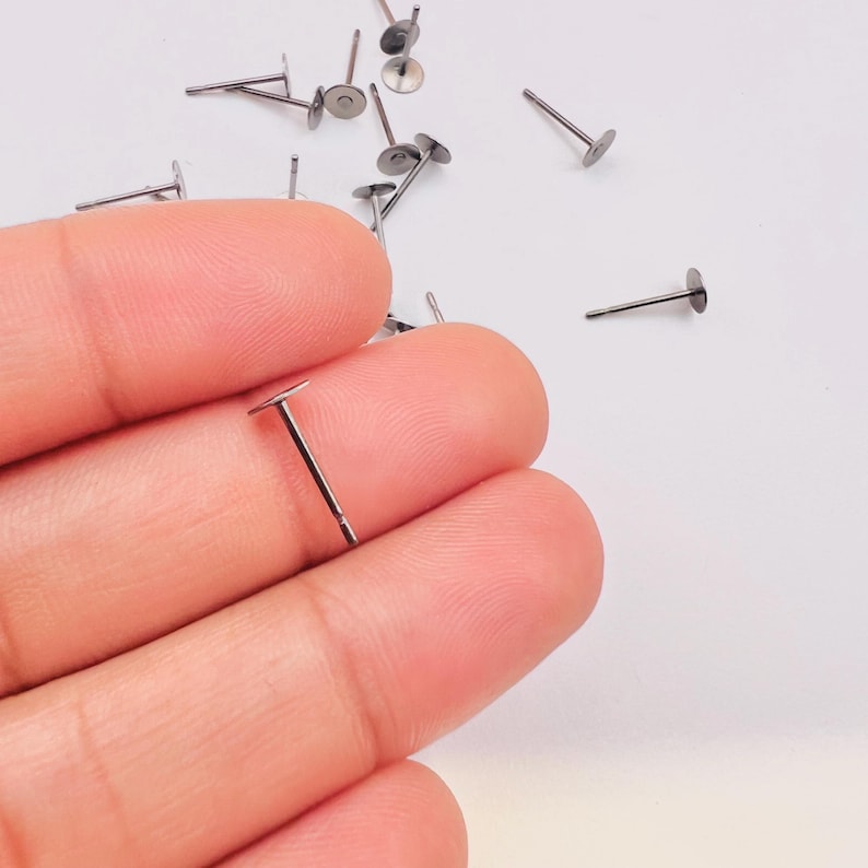 Pu&ograve; includere: Una collezione di piccoli perni per orecchini color argento con cime piatte e rotonde e perni dritti. I perni sono sparsi su una superficie bianca, uno tenuto tra le dita. Questi componenti per orecchini sono ideali per la creazione di gioielli.