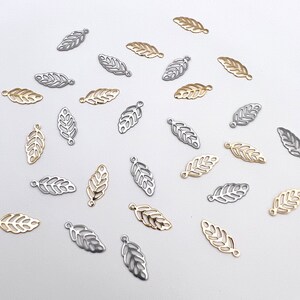 May include: A collection of gold and silver leaf charms, scattered on a white background. The charms are in the shape of leaves with a delicate, intricate design.