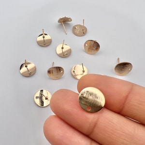May include: A close-up of ten gold-toned earring posts with round, flat backs. The posts are arranged in a scattered pattern on a white surface.