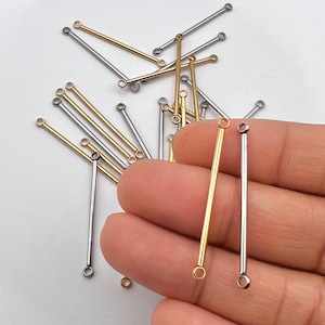 May include: Close-up of gold and silver-colored bar connectors for jewelry making. The connectors have a small loop at each end. Some connectors are held in a hand, while others are scattered on a white surface.