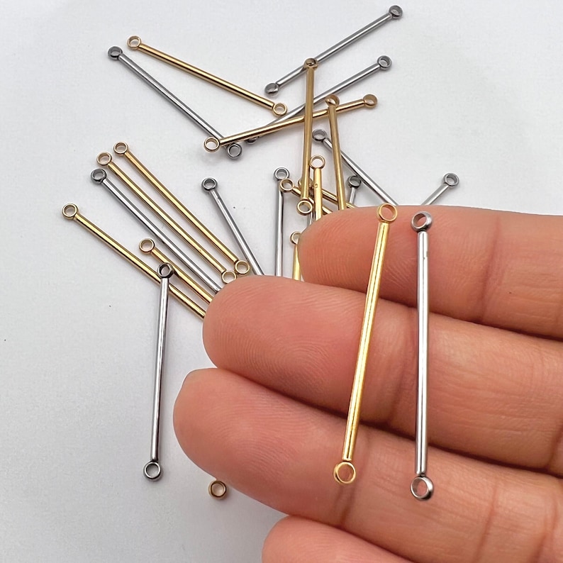 May include: Close-up of gold and silver-colored bar connectors for jewelry making. The connectors have a small loop at each end. Some connectors are held in a hand, while others are scattered on a white surface.