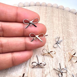 May include: A close-up of silver and gold bow charms with a jump ring. The charms are on a wooden surface.