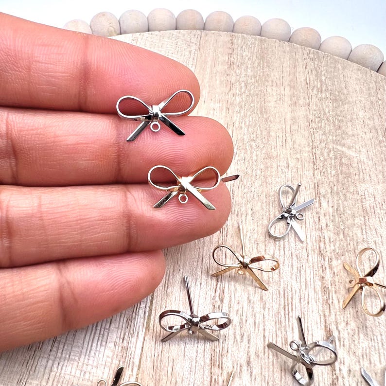 May include: A close-up of silver and gold bow charms with a jump ring. The charms are on a wooden surface.