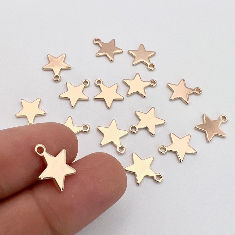 K&ouml;nnte beinhalten: Goldfarbene Metall-Stern-Anh&auml;nger mit einem Sprungring. Die Anh&auml;nger sind auf einer wei&szlig;en Oberfl&auml;che verstreut.