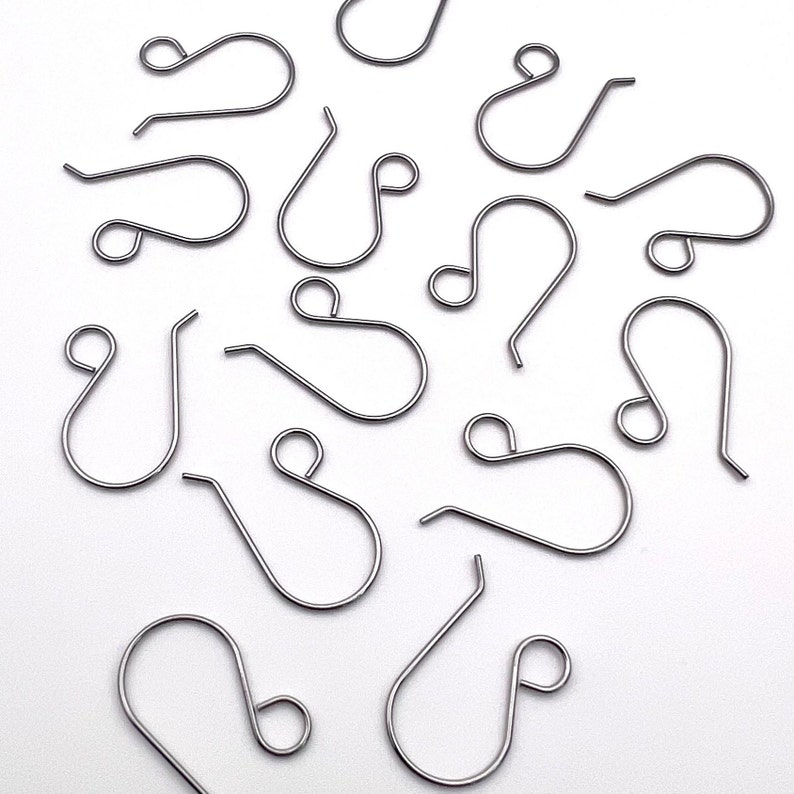 May include: A group of silver-toned earring hooks, scattered on a white surface. The hooks are all the same size and shape, with a small loop at the top and a larger loop at the bottom.