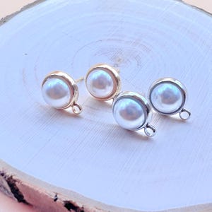 May include: Four stud earrings with a gold and silver finish. Each earring features a large, round, white pearl. The earrings are displayed on a light-colored wooden surface. The earrings have a small loop at the bottom.