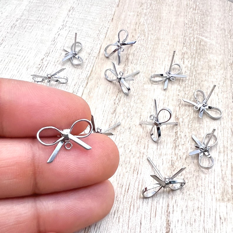 May include: A group of silver bow-shaped earring findings. The findings are small and delicate, with a loop at the bottom for attaching a dangle or bead.