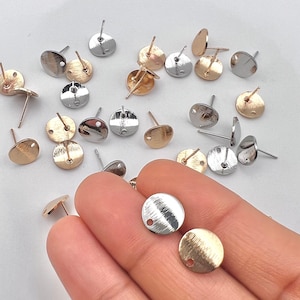 May include: Assortment of silver, gold, and rose gold earring posts with a flat round disc. The earring posts are scattered on a white surface.