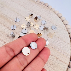 May include: A collection of round earring blanks in silver and gold tones. The blanks are flat and have a small hole in the center for attaching earring posts. The blanks are arranged on a wooden surface.