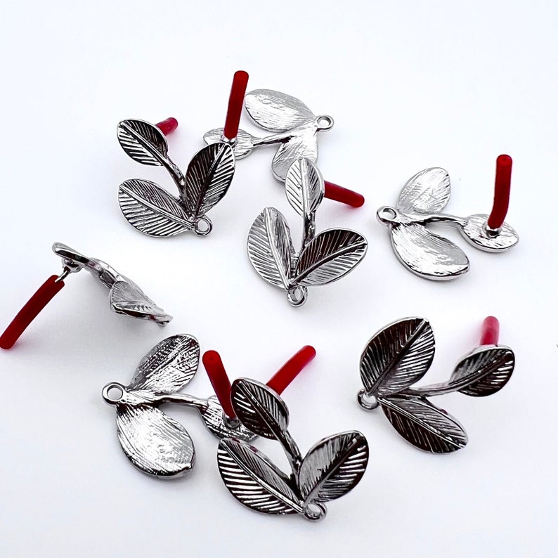 May include: Silver leaf charms with red stems. The charms are shaped like three leaves connected to a stem. The charms are perfect for jewelry making or crafting.