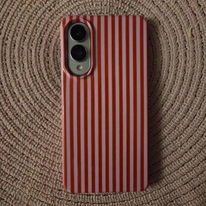 May include: A rectangular smartphone case featuring a vertical striped pattern in alternating shades of bright red and light pink. The case includes a cutout section at the top left corner for a dual camera lens and a flash component.