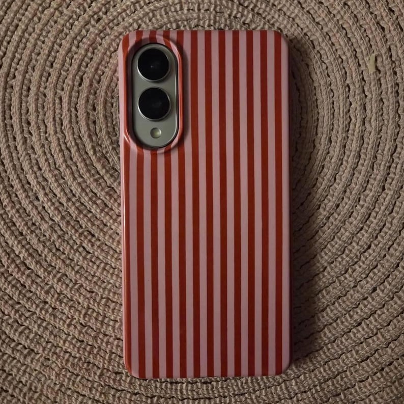 May include: A rectangular smartphone case featuring a vertical striped pattern in alternating shades of bright red and light pink. The case includes a cutout section at the top left corner for a dual camera lens and a flash component.