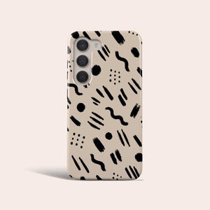 May include: A beige phone case featuring a black abstract design. The pattern includes brushstrokes, dots, and squiggly lines. The case has a camera cutout and buttons.