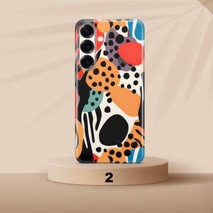 May include: A smartphone case featuring a vibrant abstract pattern of black, orange, red, and teal shapes on a cream background. The design includes various organic blobs and black polka dots, with a triple-lens camera cutout on the upper left side.