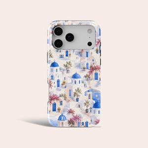 May include: A smartphone case featuring a watercolor-style illustration of a Mediterranean village with white buildings, blue domed roofs, blue doors, and vibrant pink flowering bushes. The design covers the back of the phone case entirely.