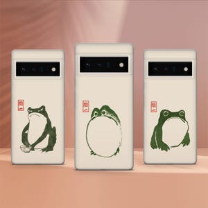 May include: Three white phone cases with different green frog illustrations. The cases have a black camera lens and the word "閒" in red on the top left corner.