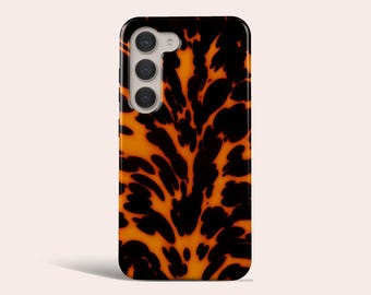 May include: A rectangular phone case with a tortoiseshell pattern in shades of black and orange. The case has a camera module with three lenses. The design is a repeating pattern of dark, irregular shapes against a lighter background.