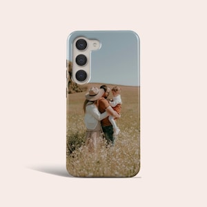 Puede incluir: Funda de teléfono con un retrato familiar en un campo de flores silvestres. La imagen muestra a una pareja abrazándose, sosteniendo a un niño. La funda tiene un borde azul claro y un recorte para la cámara.