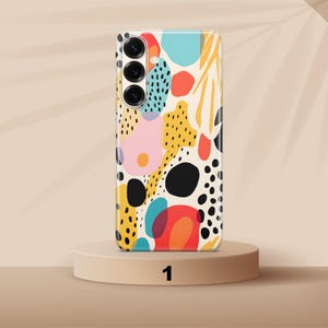 May include: A rectangular smartphone case featuring a cream-colored background with an abstract pattern of black, red, yellow, pink, and teal shapes, including solid circles, organic blobs, and clusters of small black dots, with a triple camera cutout.