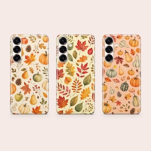 May include: Three smartphone cases featuring watercolor illustrations. The left case has pumpkins, pears, and leaves on a cream background. The center case displays various autumn leaves. The right case shows pumpkins, gourds, and mushrooms on a peach background.